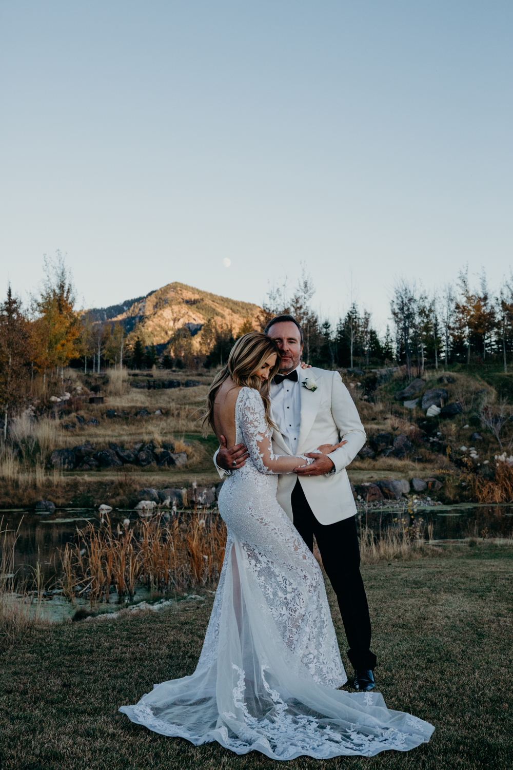 A bride wearing a beautiful lace Berta gown embraces groom as moon rises in Jackson Hole, Wyoming