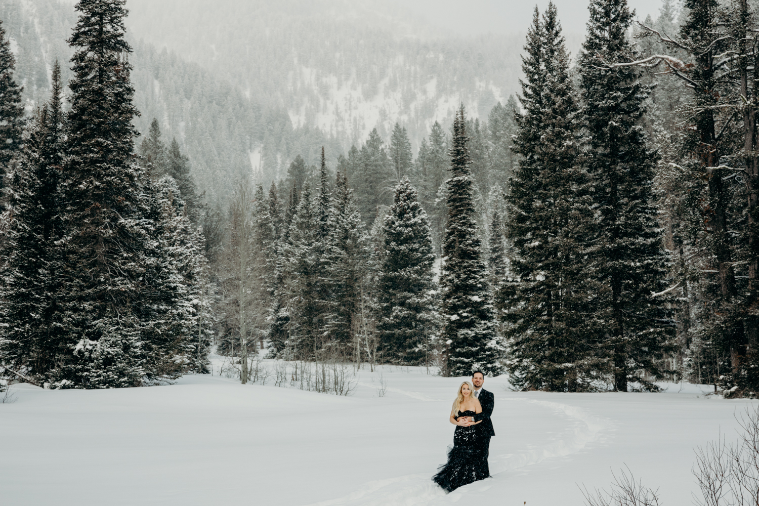Man in black suit holds woman in black gown amongst snowy trees in Teton Pass during Jackson Hole engagement photos
