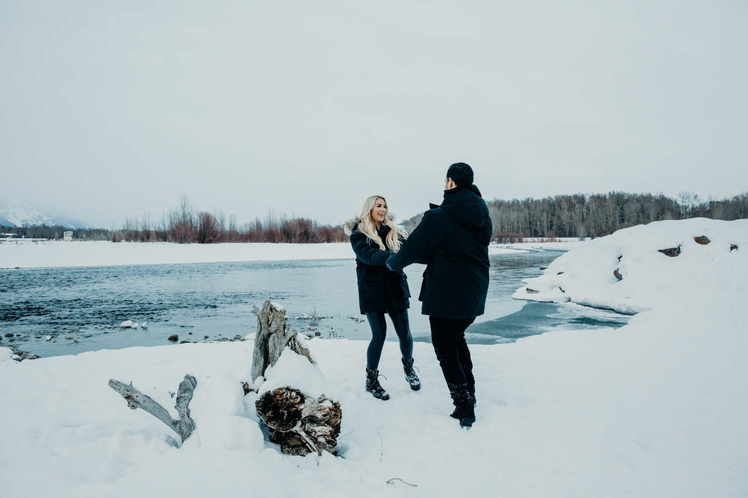 A couple wearing black parkas swing eachother smiling on the edge of the Snake River in Jackson Hole Wyoming during their engagement photos