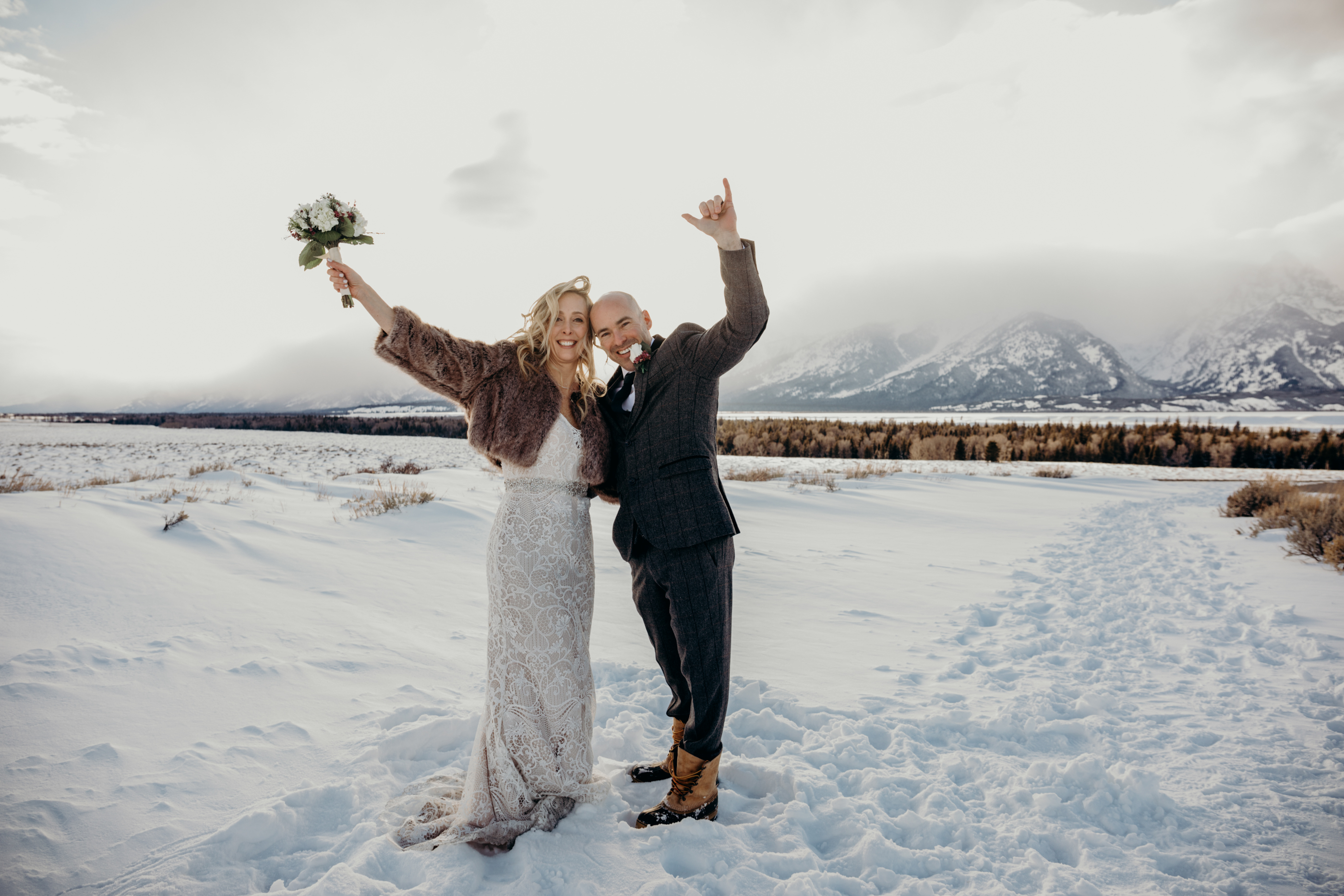 Newly married couple wearing suit, gown and fur coat raise their arms in the air in celebration during their elopement at Grand Teton National Park